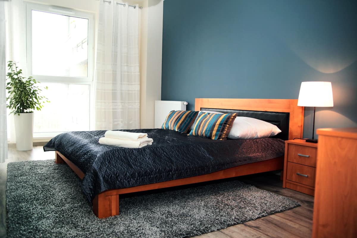A bedroom with a bed, a bedside table, a lamp, a potted plant, and a window with curtains.