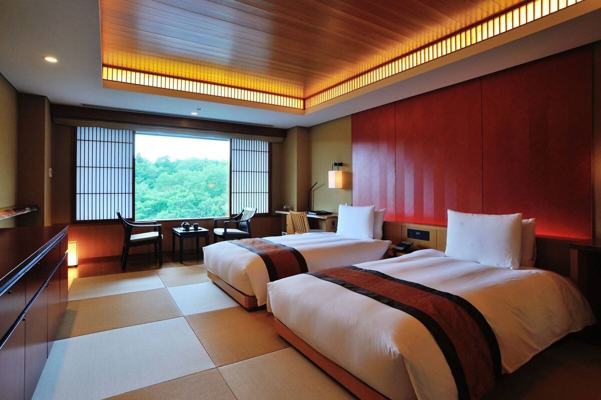 A hotel room with two beds, a desk with chairs, a view of greenery, and a red accent wall.