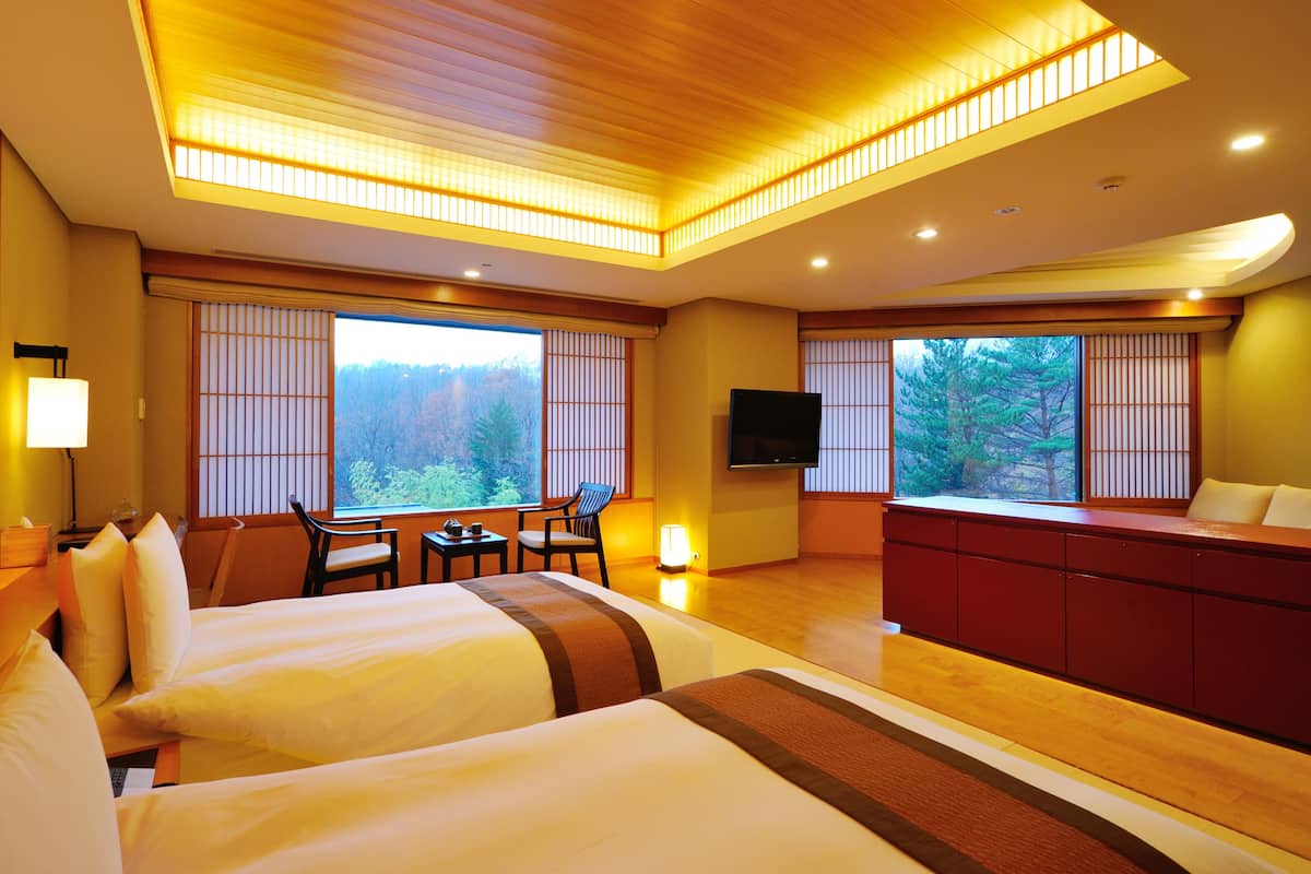 A hotel room with two beds, a TV, a desk with a chair, and large windows with a forest view.