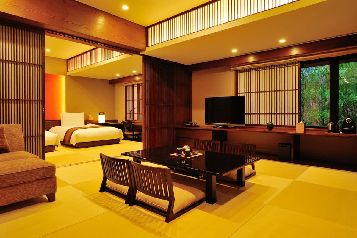 A hotel room with a bed, a TV, a dining table with chairs, and a view of greenery outside.