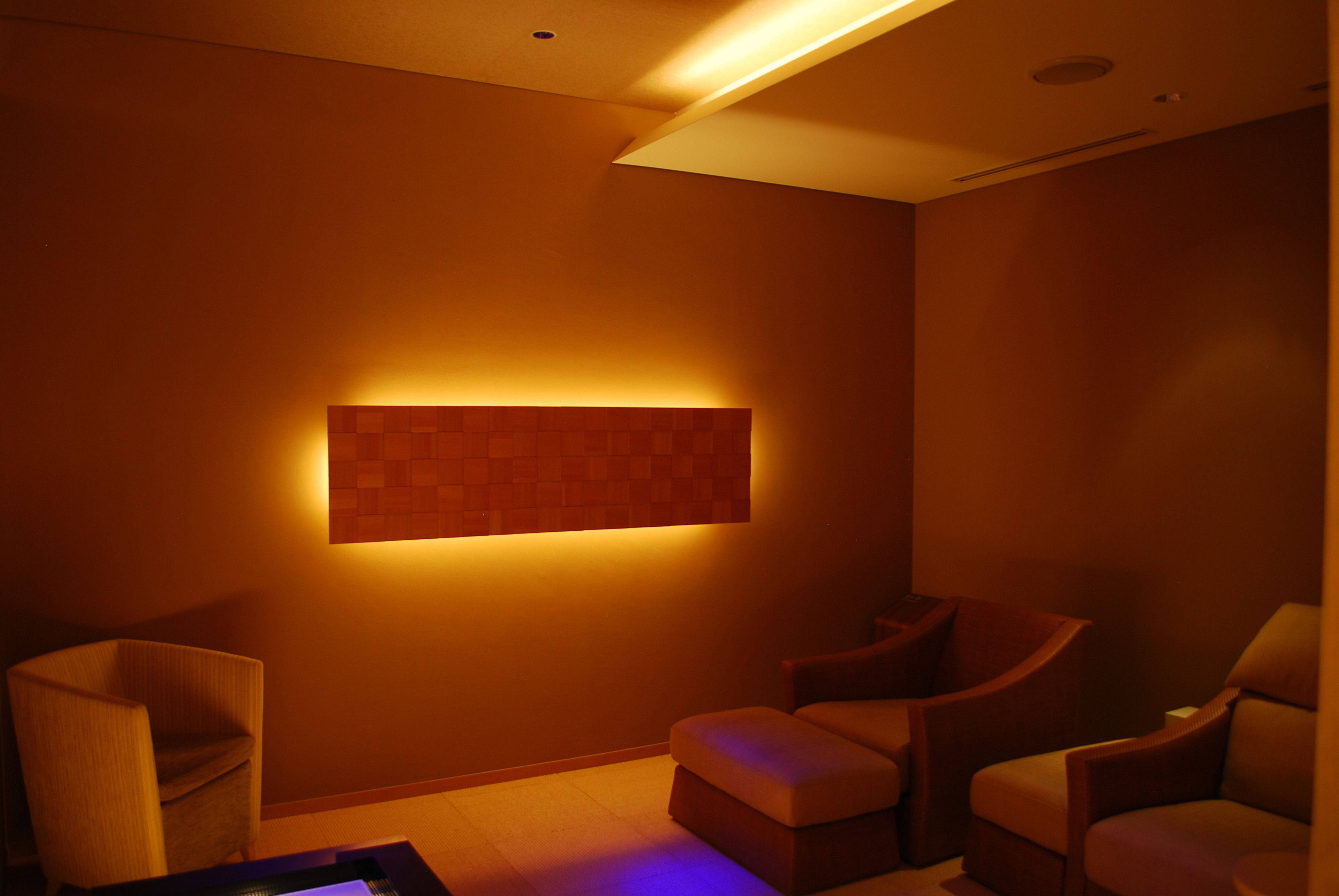 A dimly lit room with a modern design, featuring a wall-mounted light panel and two cushioned chairs.