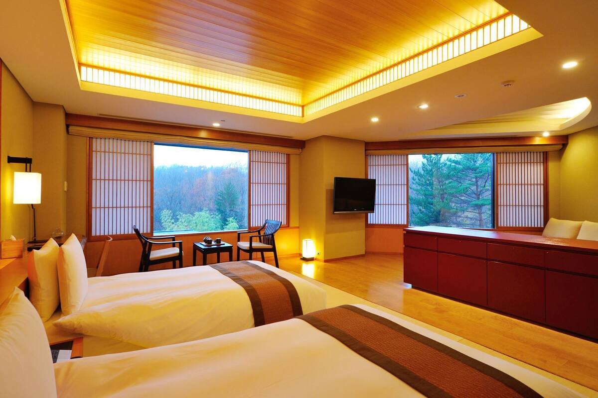 A hotel room with two beds, a TV, a dining area, and large windows with a forest view.