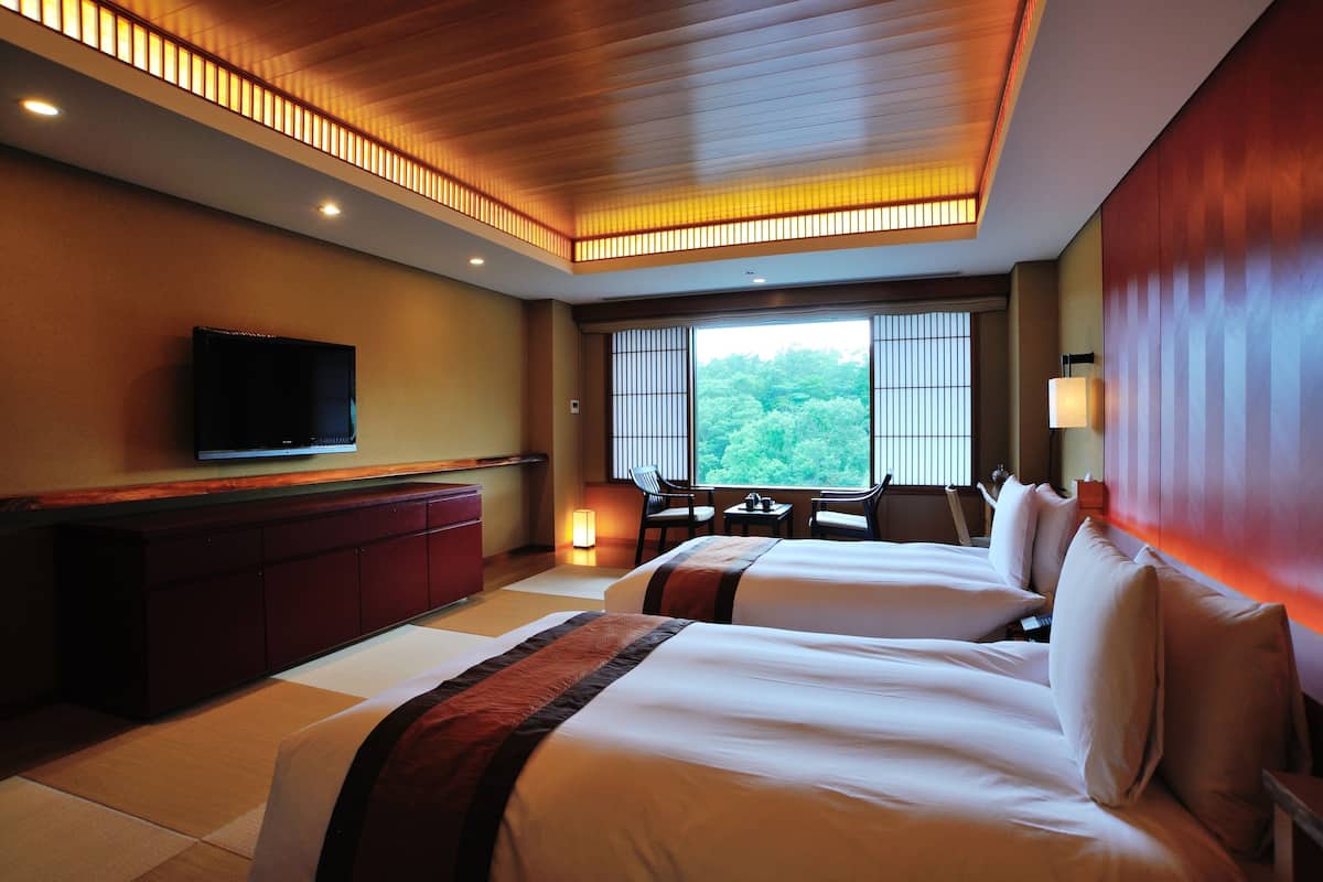 A hotel room with two beds, a TV, a desk, and a view of trees through the window.