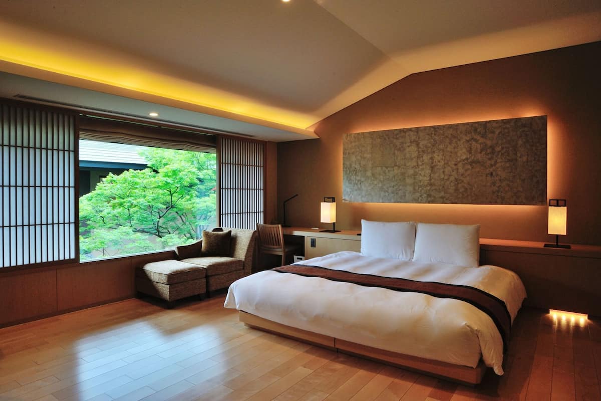 A modern hotel room with a large bed, a seating area, and a view of greenery outside.