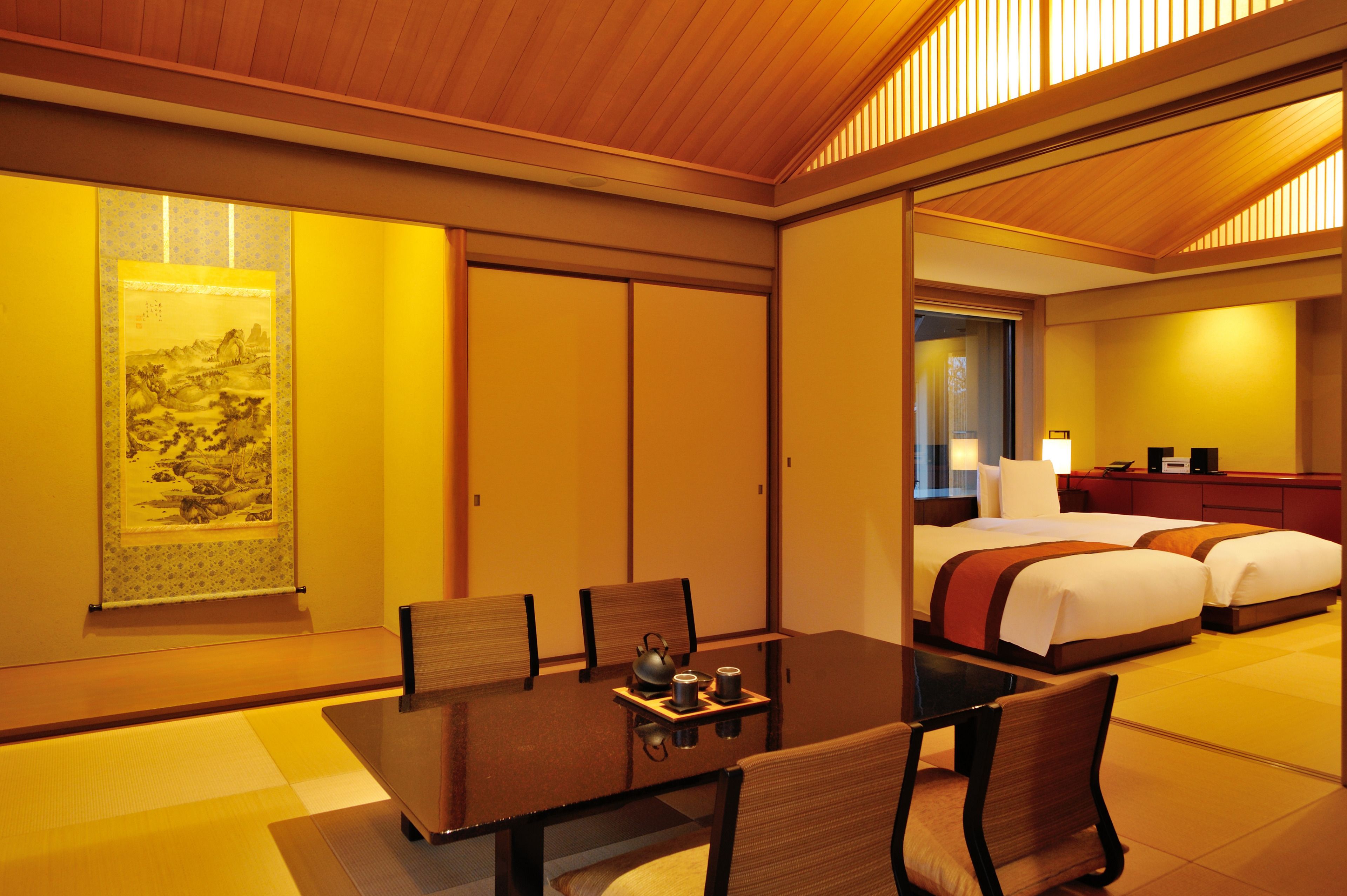 A hotel room with a traditional Japanese aesthetic, featuring tatami flooring, a low table with a tea set, and a bed with a red accent.