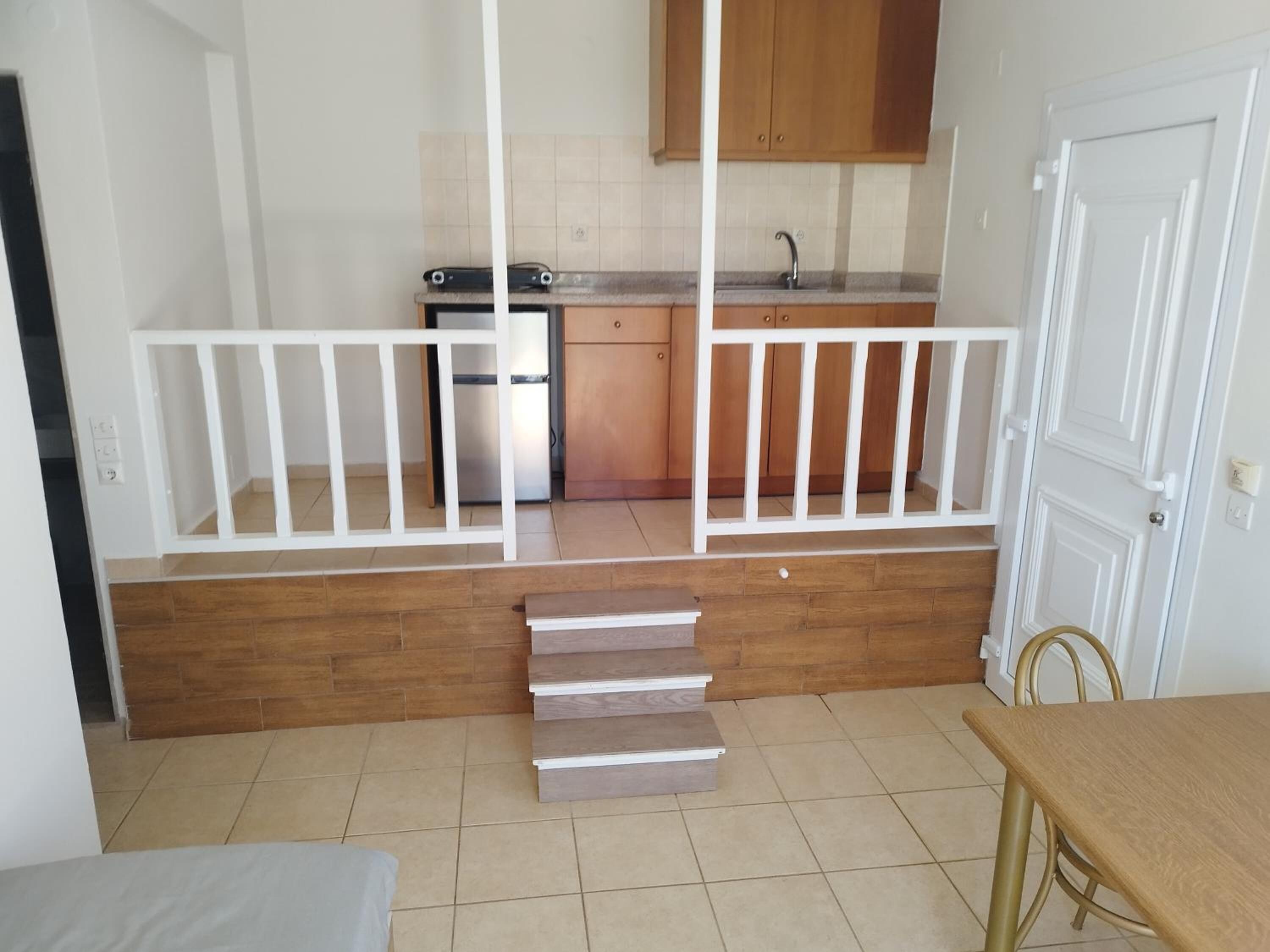 Eine kleine, offene Küche mit Kühlschrank, Herd und Holzschränken. In der Mitte eine weiße Geländer und im Vordergrund ein kleiner Tisch mit einem Stuhl.