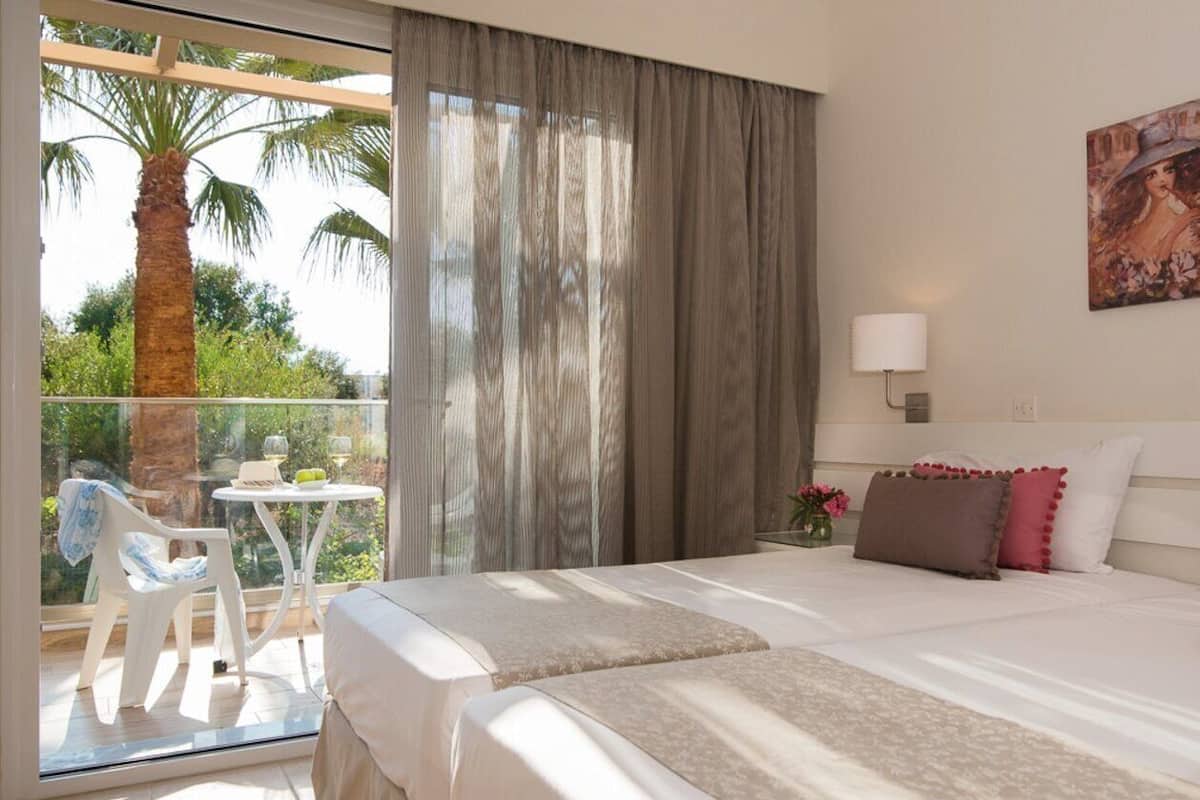 Habitación de hotel con balcón, cama con ropa blanca, una pequeña mesa con una silla y un cuadro en la pared.