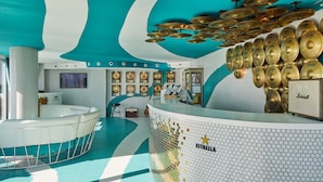 A modern interior with a teal and white color scheme, a central counter with a Marshall amplifier, and a wall decorated with numerous golden cymbals.