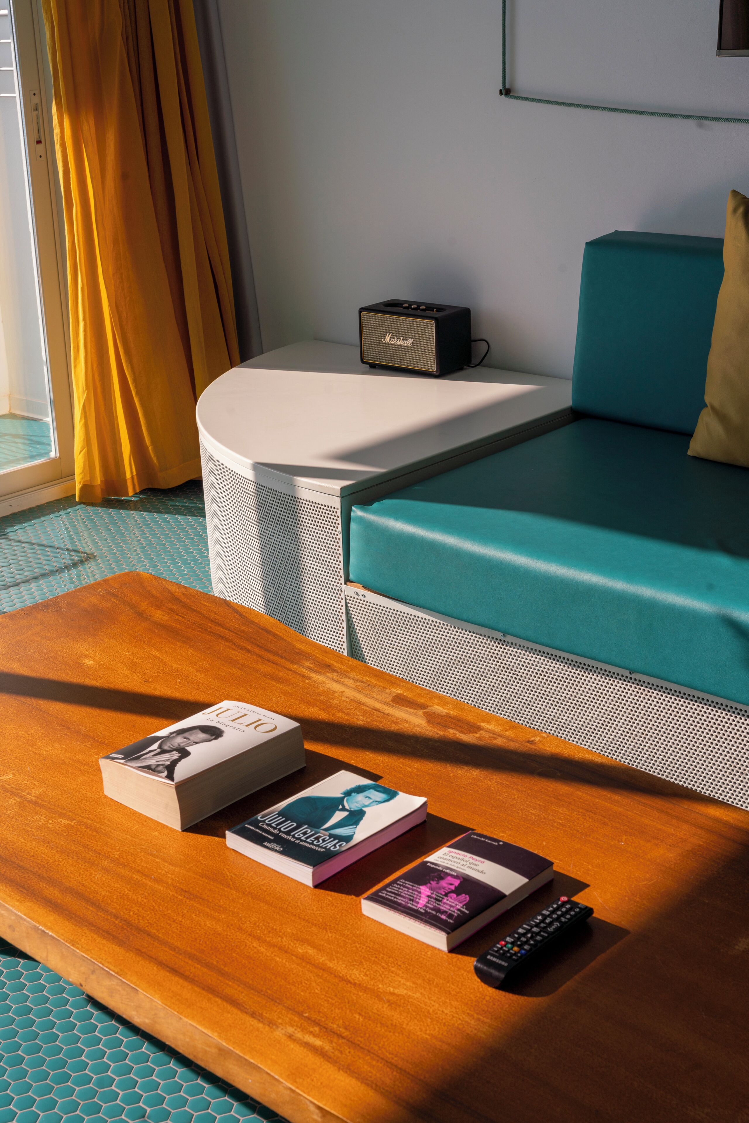 Une pièce avec une table basse en bois, un pouf rond blanc, un canapé turquoise, un haut-parleur Marshall et une fenêtre avec des rideaux jaunes.