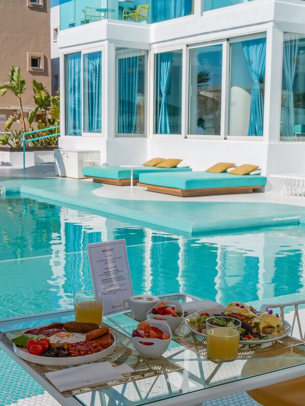 A poolside breakfast setup with a menu sign, a glass of orange juice, and plates of food including eggs, beans, and toast.