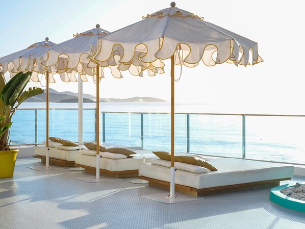 A sunlit outdoor seating area with white cushioned loungers and decorative umbrellas, overlooking a body of water.