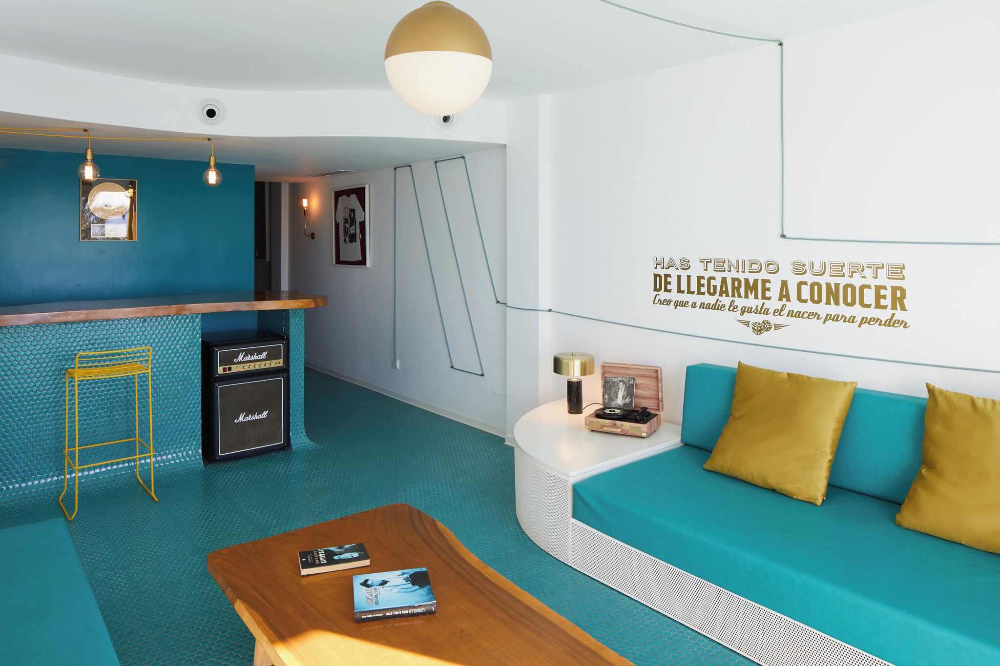 Una habitación de hotel moderna con una decoración azul aguamarina, una zona de bar con un amplificador Marshall, un sofá con cojines amarillos y una pared con una cita motivacional.