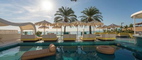 A pool area with lounge chairs, umbrellas, and palm trees.