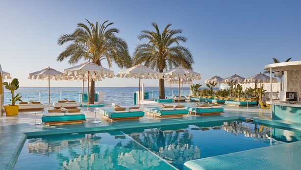A pool area with lounge chairs and umbrellas, palm trees, and a view of the sea.