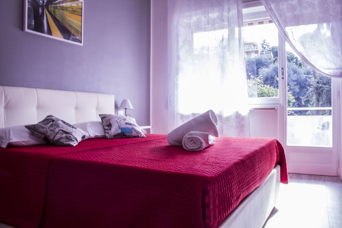 A neatly made bed with a red blanket, white pillows, and a rolled towel.
