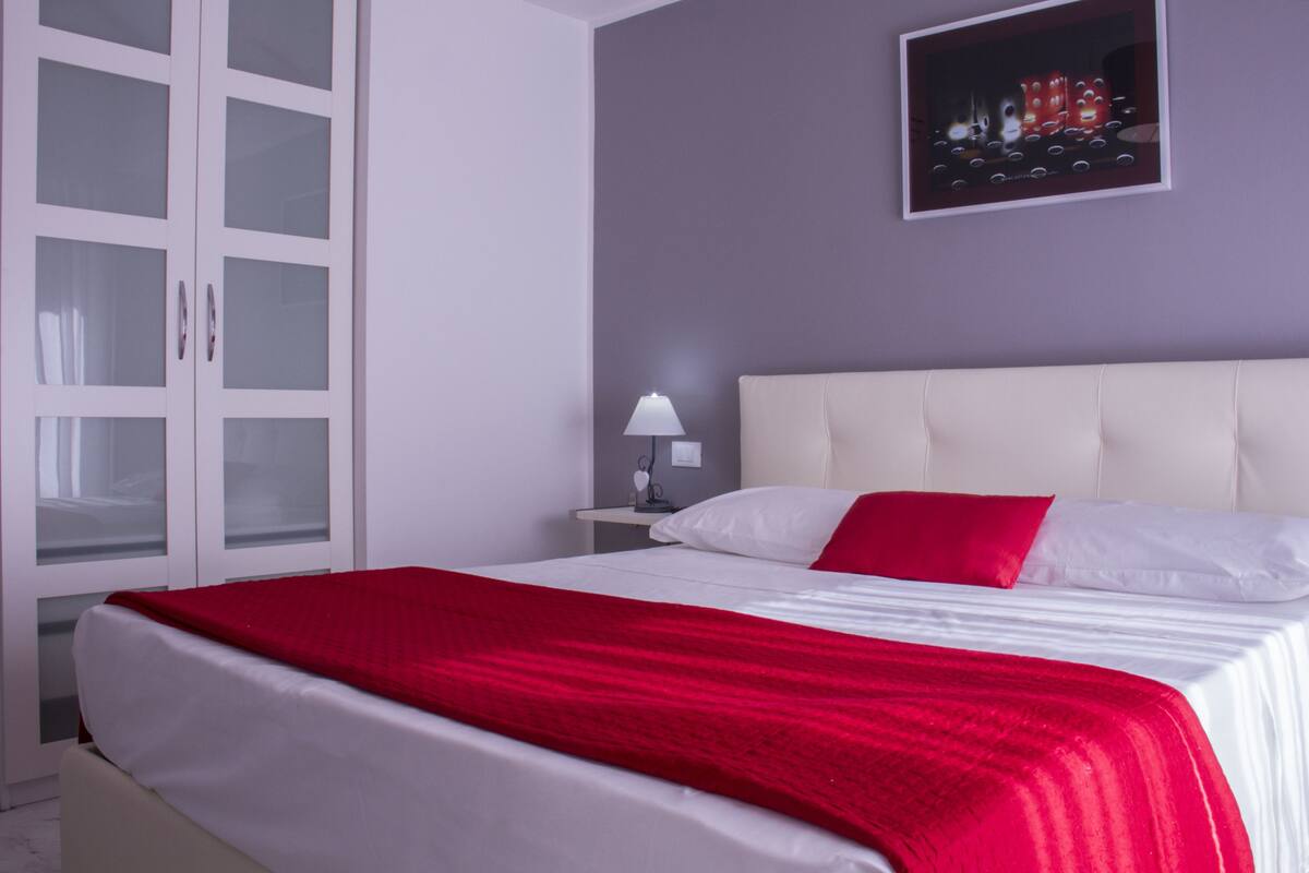 A bedroom with a bed, white headboard, red and white bedding, a nightstand with a lamp, and a wardrobe with glass doors.