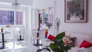 A bright, modern living room with a white sofa, red cushions, and a large plant. There is a dining area with white chairs and a table, and a window with a view of buildings outside. - Domus Raffaello suite in Rome (Rome)