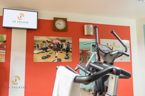 Zona de gimnasio con equipo de ejercicio, un reloj y un televisor que muestra el logotipo del hotel.
