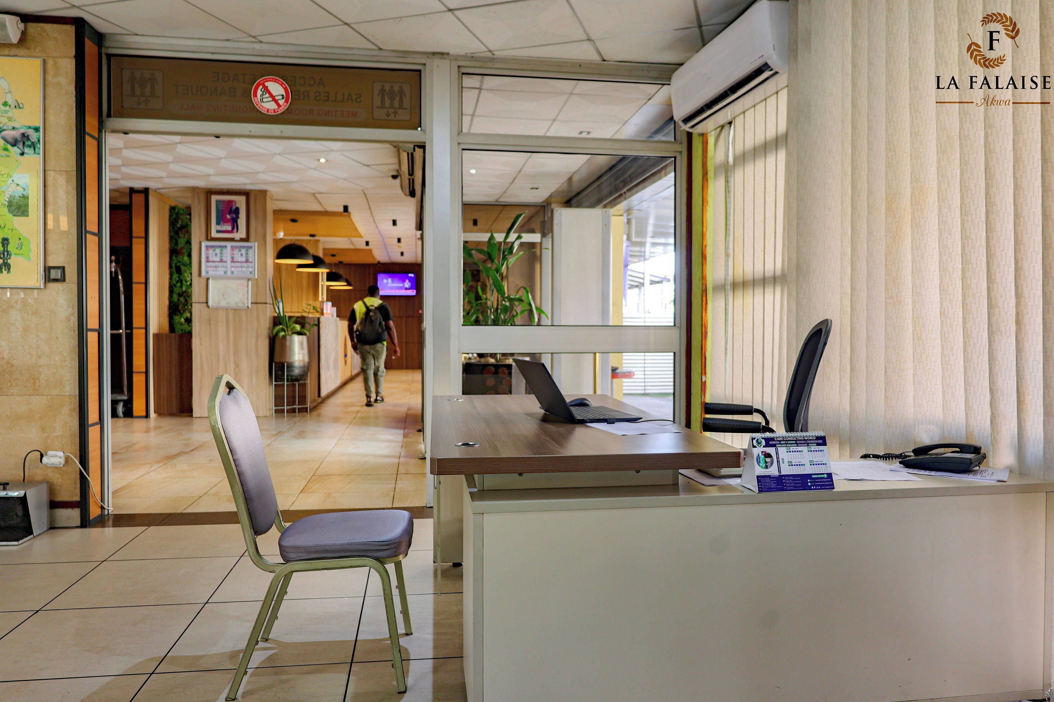 A reception desk with a computer, a telephone, and a calendar. There is a chair in front of the desk, and a person is visible in the background.