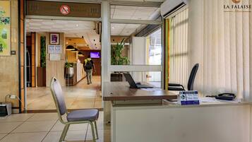 A reception desk with a computer, a telephone, and a calendar. There is a chair in front of the desk, and a person is visible in the background.
