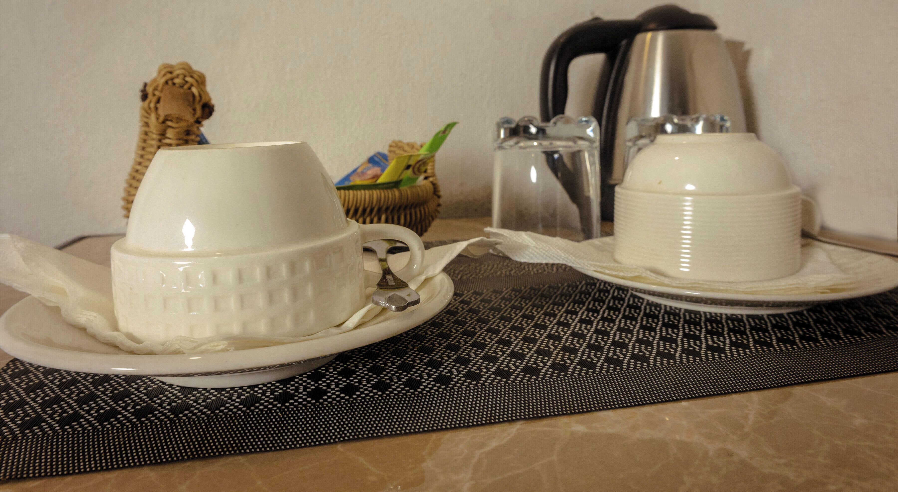 A table with two white cups on saucers, a basket with a green item, a kettle, and a glass container.