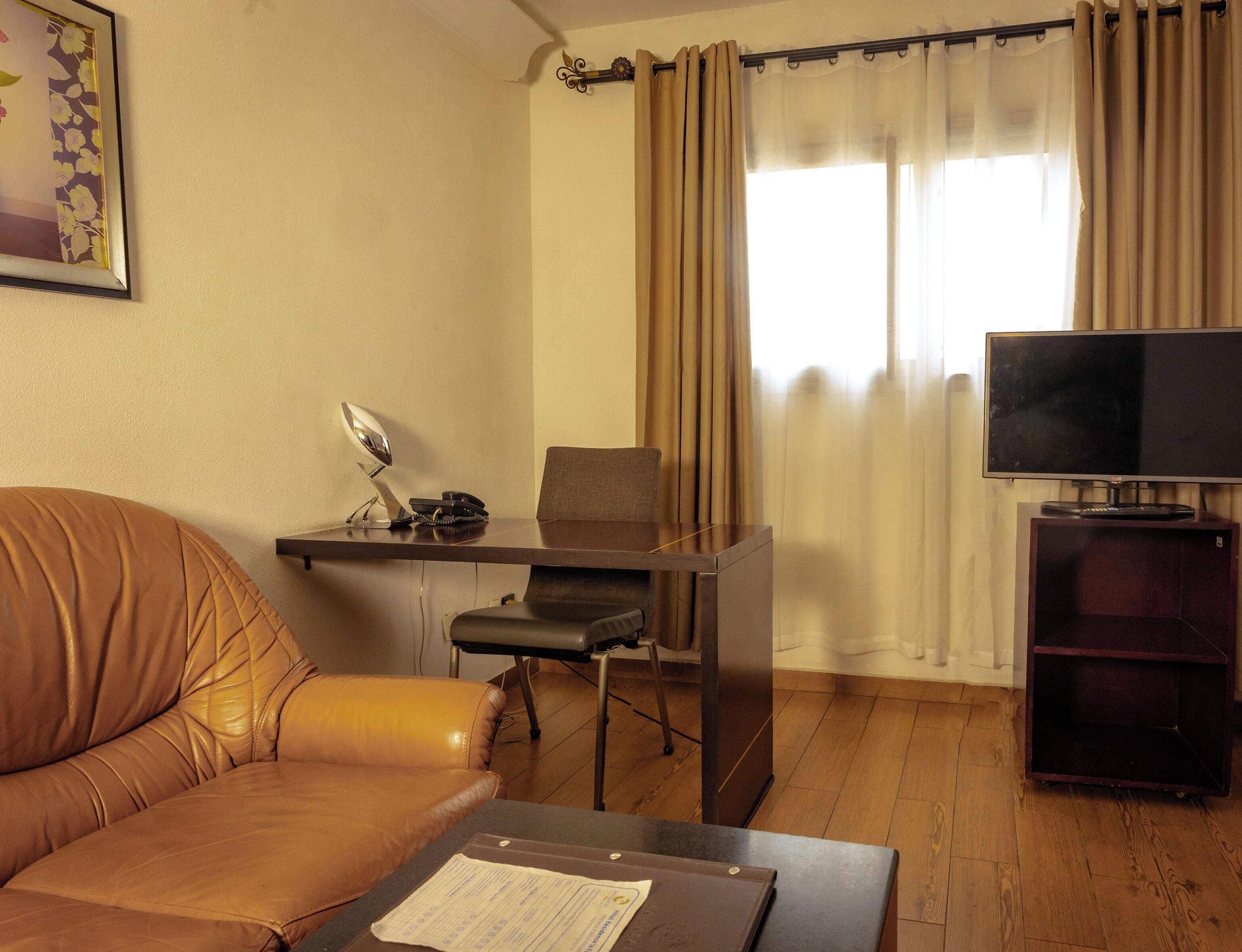 Un salon avec un canapé en cuir, un bureau avec une chaise, un téléviseur et une fenêtre avec des rideaux.