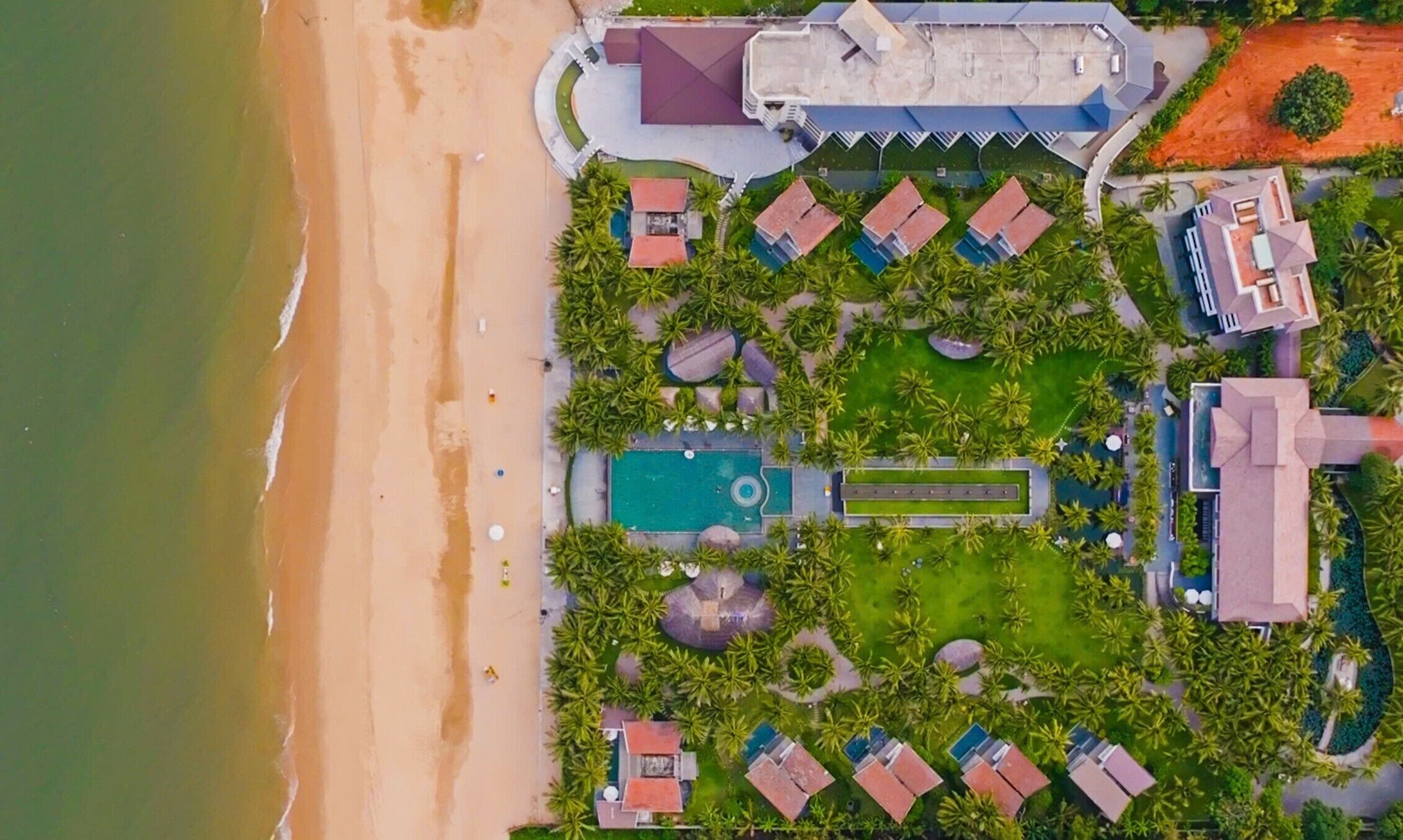 Vista aérea de un resort con playa, piscinas y varias edificaciones con techos rojos.