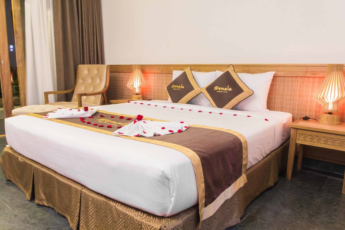 A hotel room with a neatly made bed, a chair, a bedside table with a lamp, and a headboard with decorative pillows.