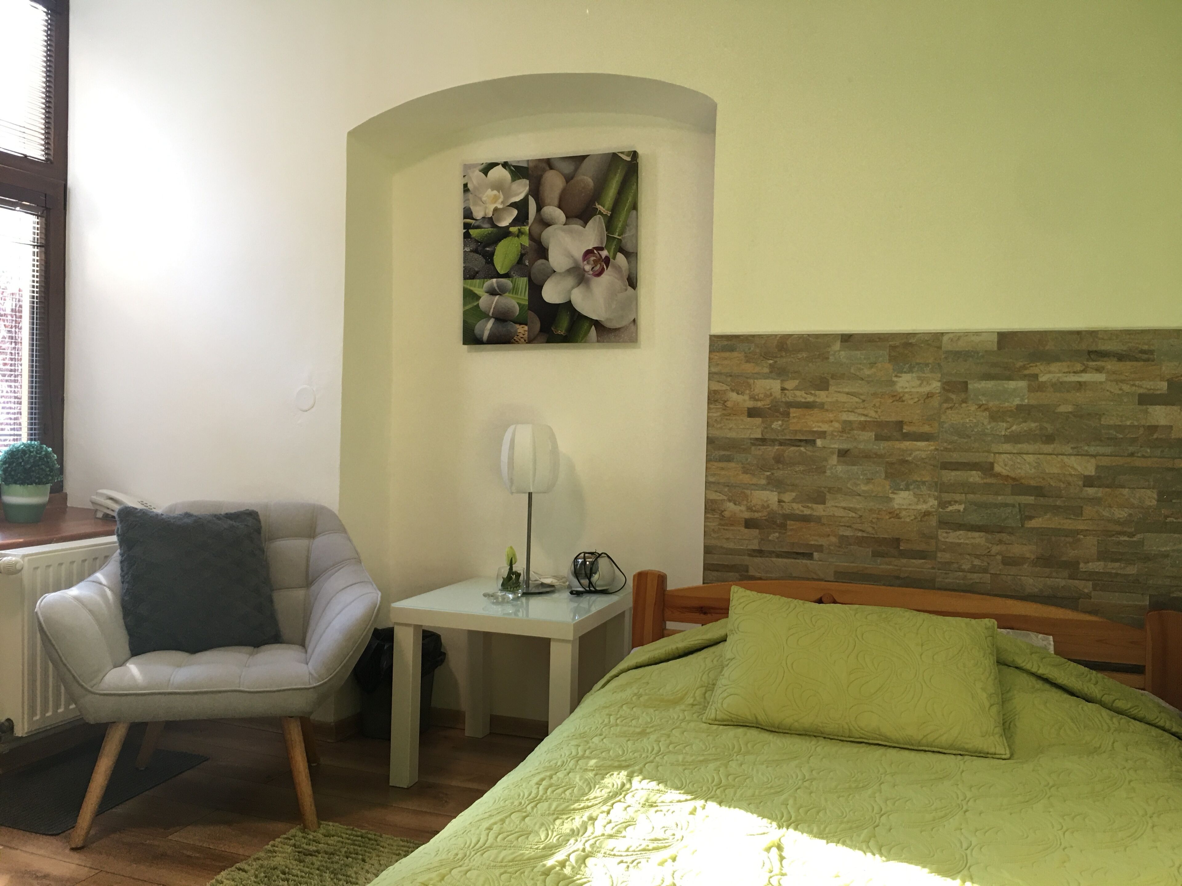 A bedroom with a green bed, a chair, a small table with a lamp, and a wall-mounted picture of flowers.