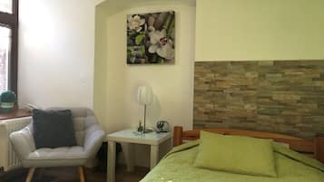 A bedroom with a green bed, a chair, a small table with a lamp, and a wall-mounted picture of flowers.