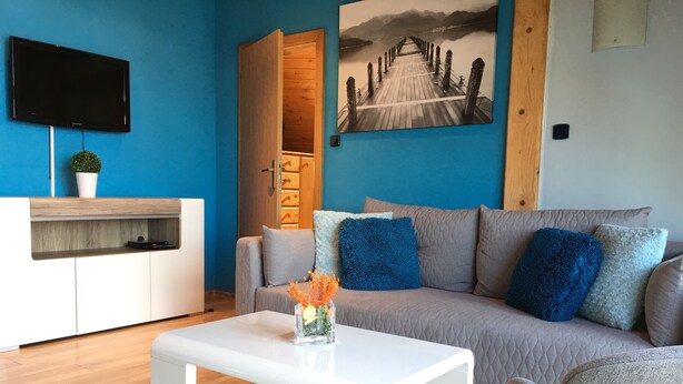 A modern living room with a white coffee table, grey sofa with blue and white cushions, a flat-screen TV mounted on a blue wall, and a wooden door leading to a kitchen. - Penzión Hradby (Prešov)