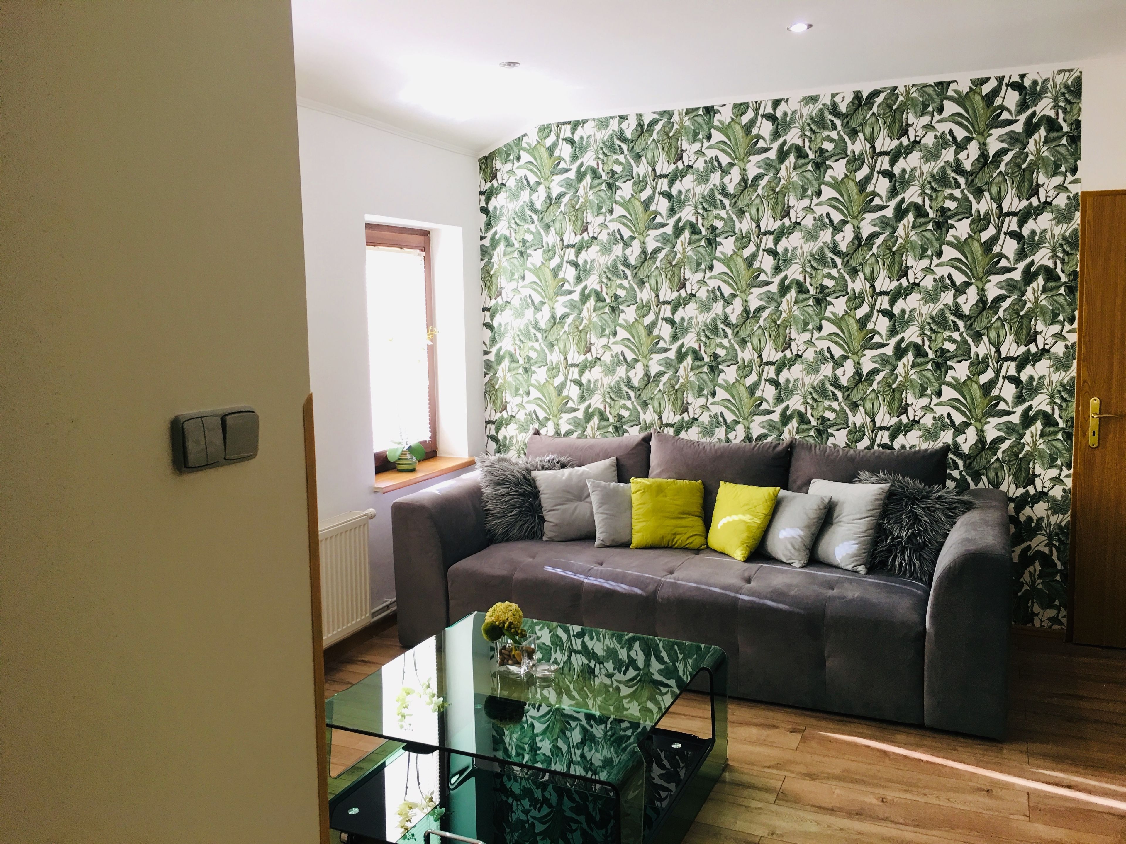 A living room with a grey sofa, a glass coffee table, and a window with curtains.