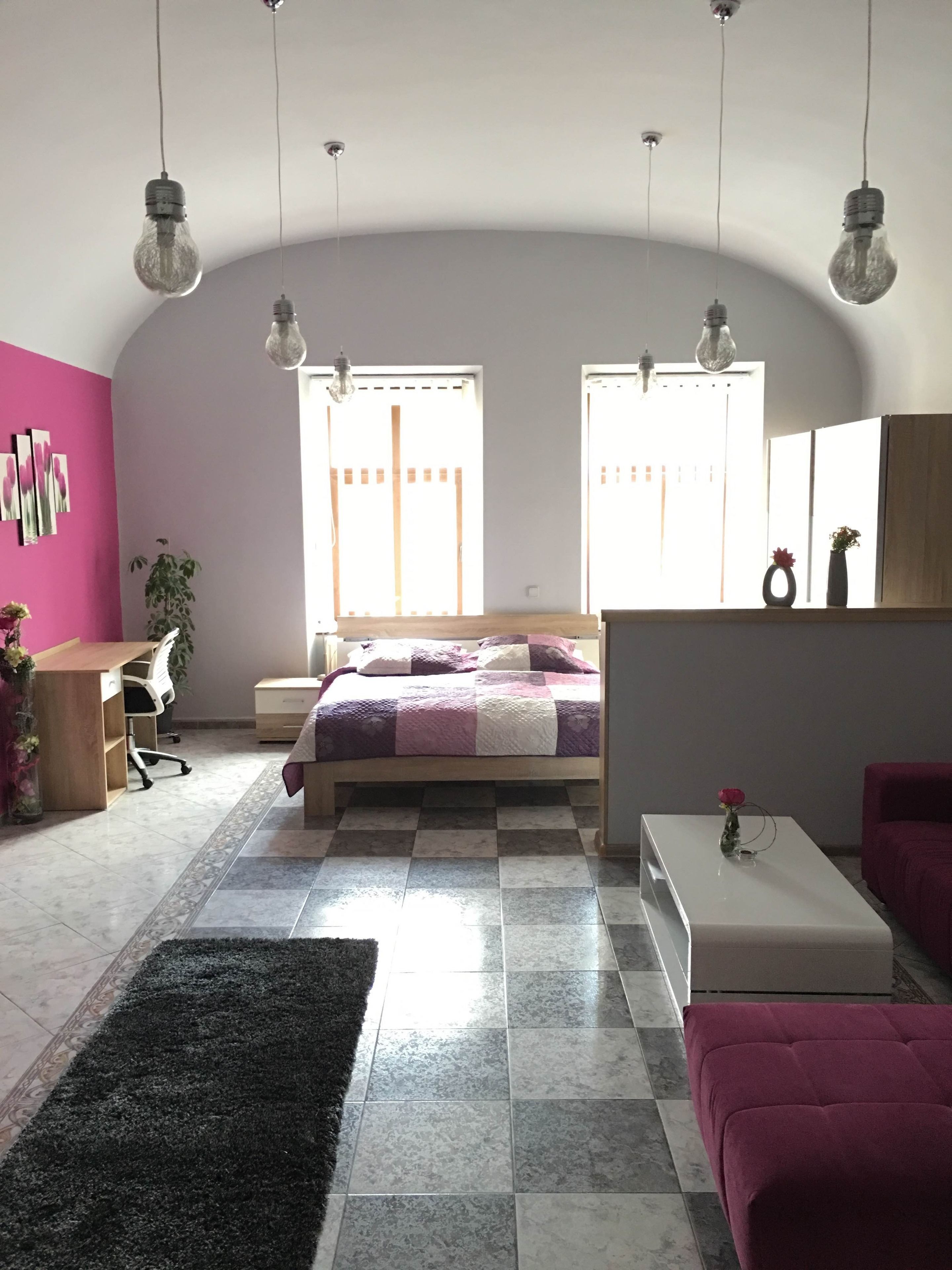Une chambre moderne avec un lit, un bureau, un canapé et une table basse. Il y a deux fenêtres avec des rideaux, une plante et un vase rempli de fleurs.