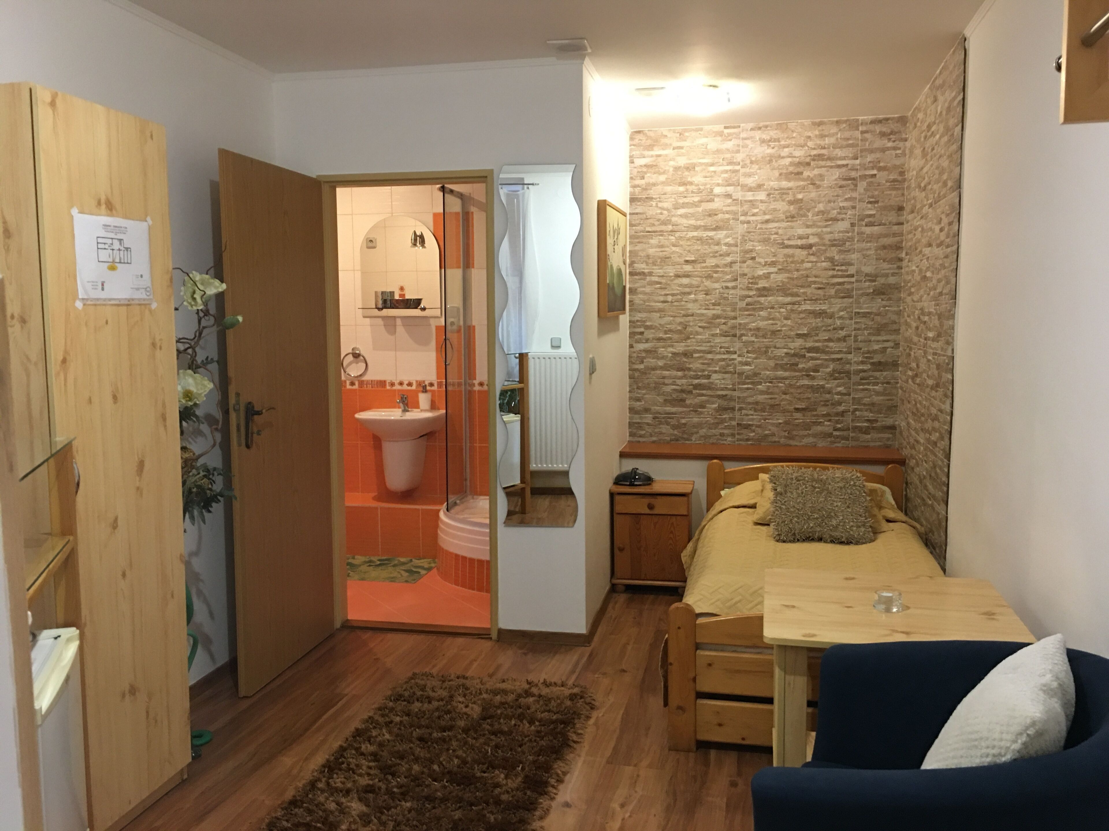 Habitació compacta amb un armari de fusta, una petita taula de nit, un llit amb capçalera blava, una taula auxiliar de fusta, una taula petita i una catifa marró.