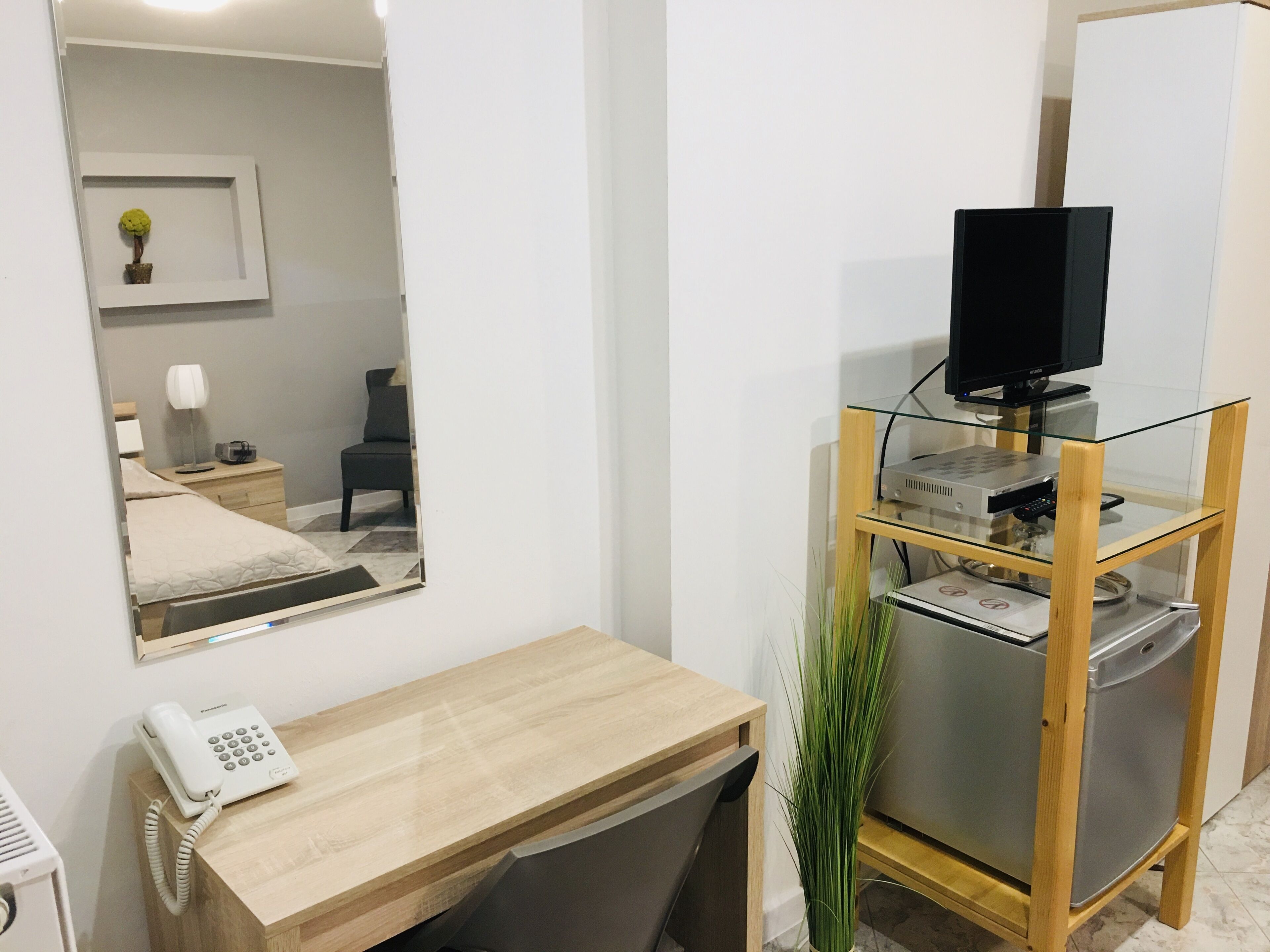 A hotel room with a wooden desk, a telephone, a television on a glass shelf, and a mirror reflecting the room.