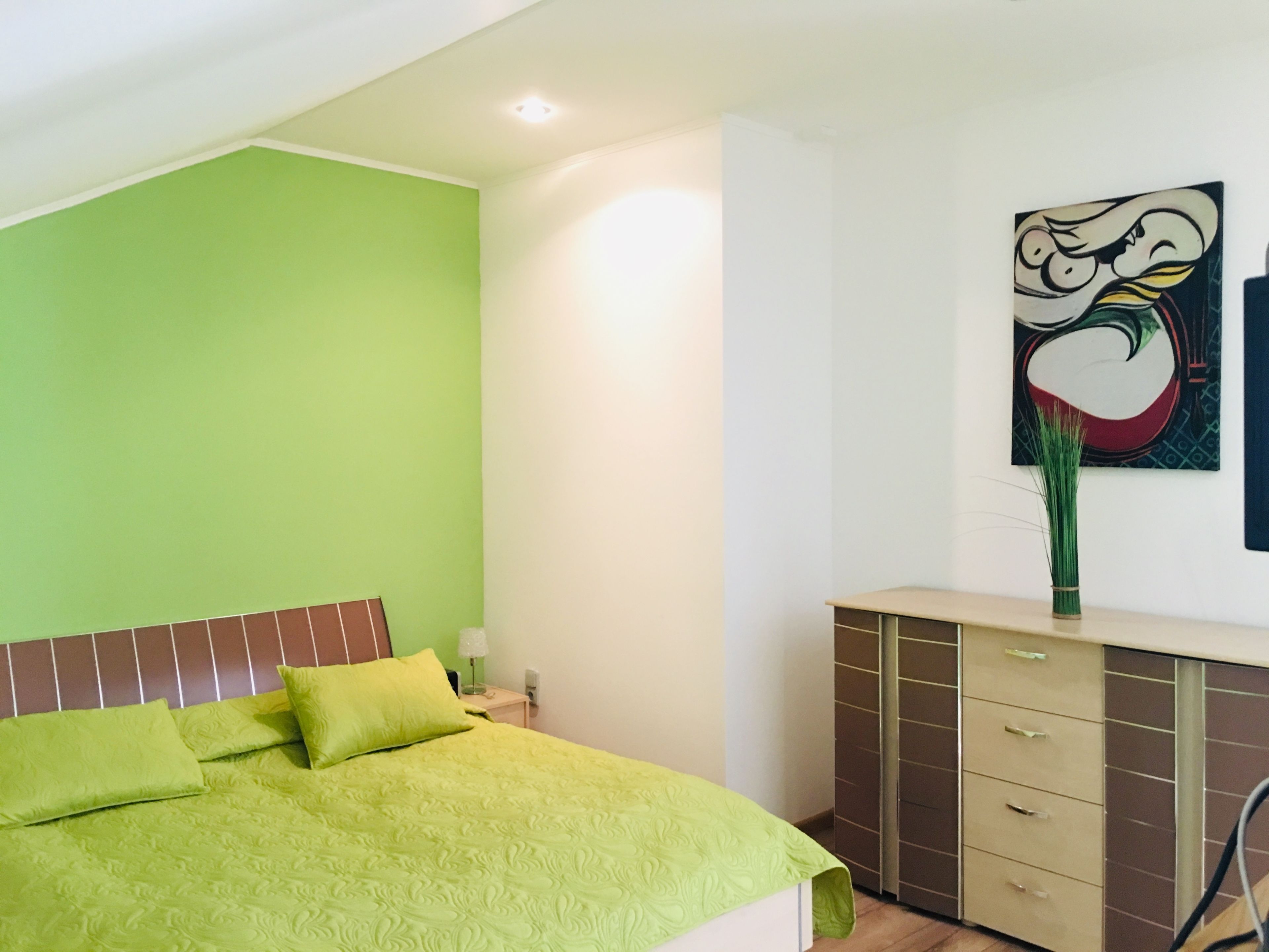 A bedroom with a green wall, a bed with green bedding, a bedside table, a lamp, a dresser, and a wall-mounted artwork.
