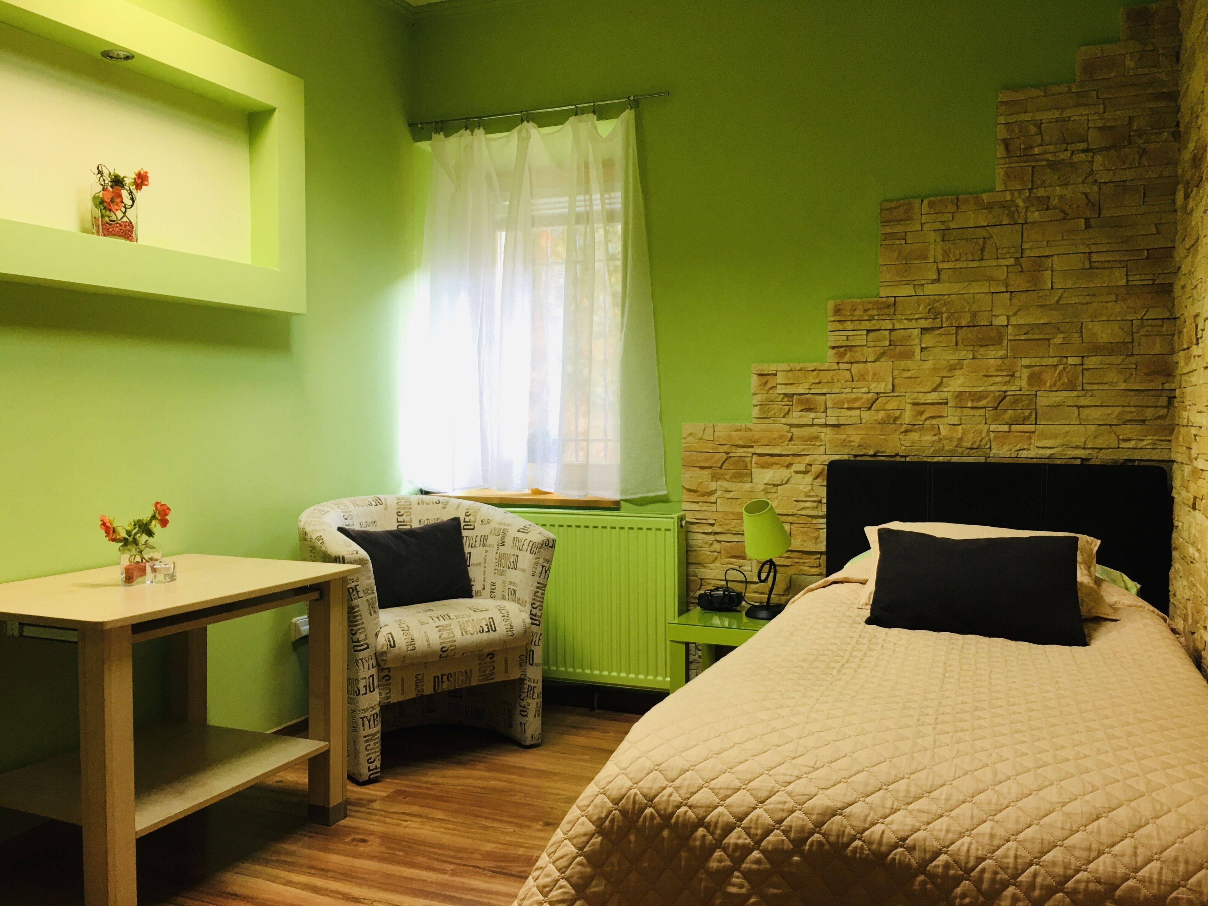 Una habitación con cama, una silla, una mesa, un ventanal con cortinas y una pared de piedra.