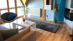A modern living room with a sloped ceiling, wooden flooring, a white sofa with a blue cushion, a white coffee table, a grey rug, a potted plant, a radiator, and a mounted TV. - Penzión Hradby (Prešov)
