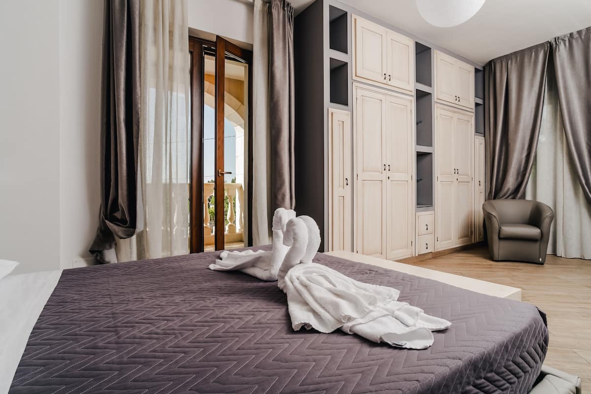 A neatly made bed with a herringbone-patterned bedspread, a pair of white towels, and a chair in a modern room with a large wardrobe and a balcony view.
