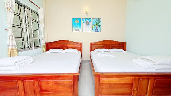 A hotel room with two wooden beds, white linens, a window with a curtain, and a wall-mounted light fixture.