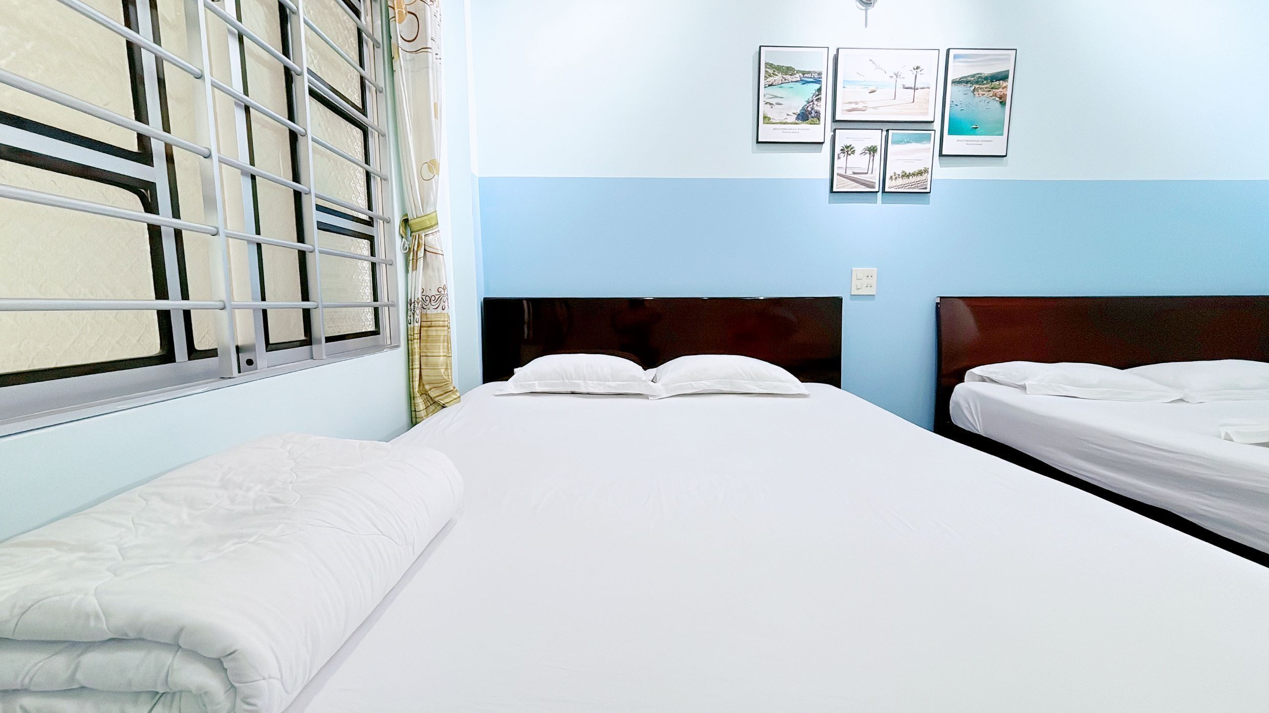Une chambre d’hôtel avec deux lits, une fenêtre avec des barreaux et un mur orné de tableaux.