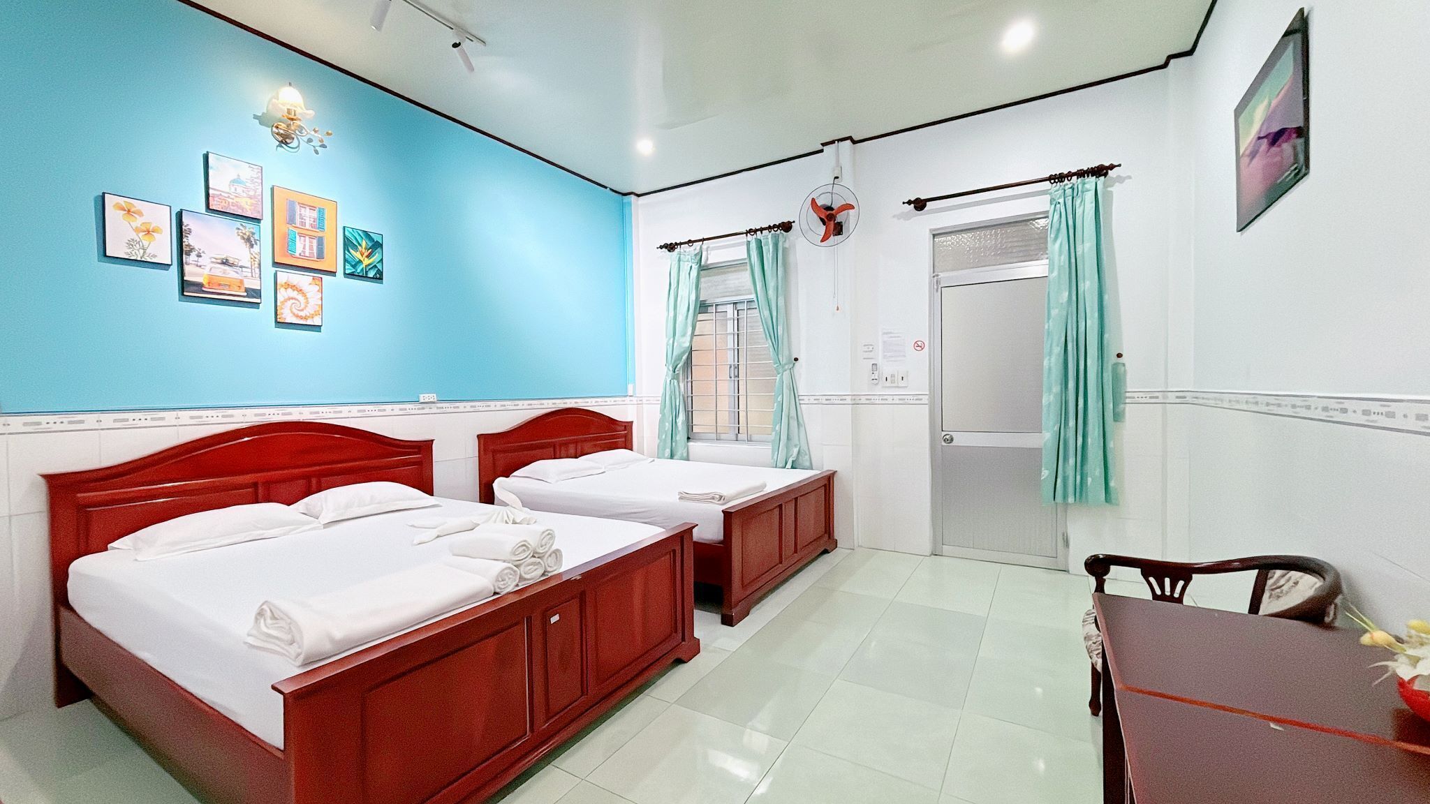 A hotel room with two wooden beds, a desk, and a chair. There are wall-mounted lights and a window with curtains.