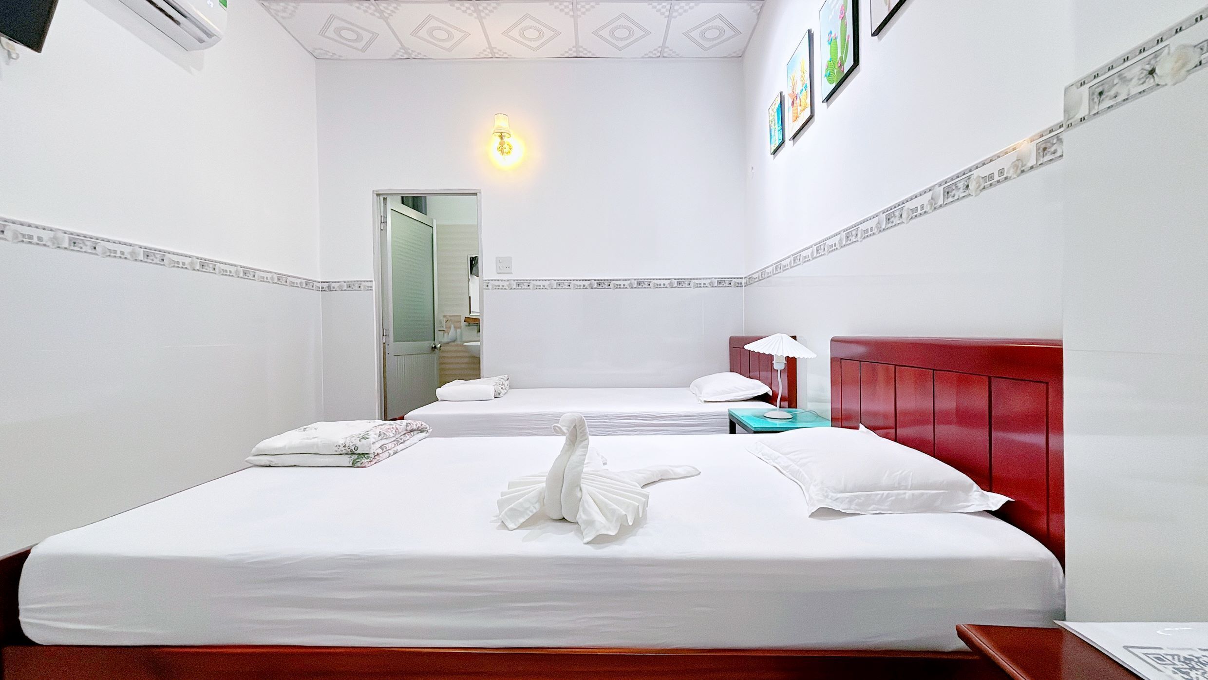 A hotel room with two beds, a red headboard, a nightstand, and a wall-mounted lamp.