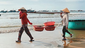 Two people walking on a beach, carrying baskets, with boats in the background.