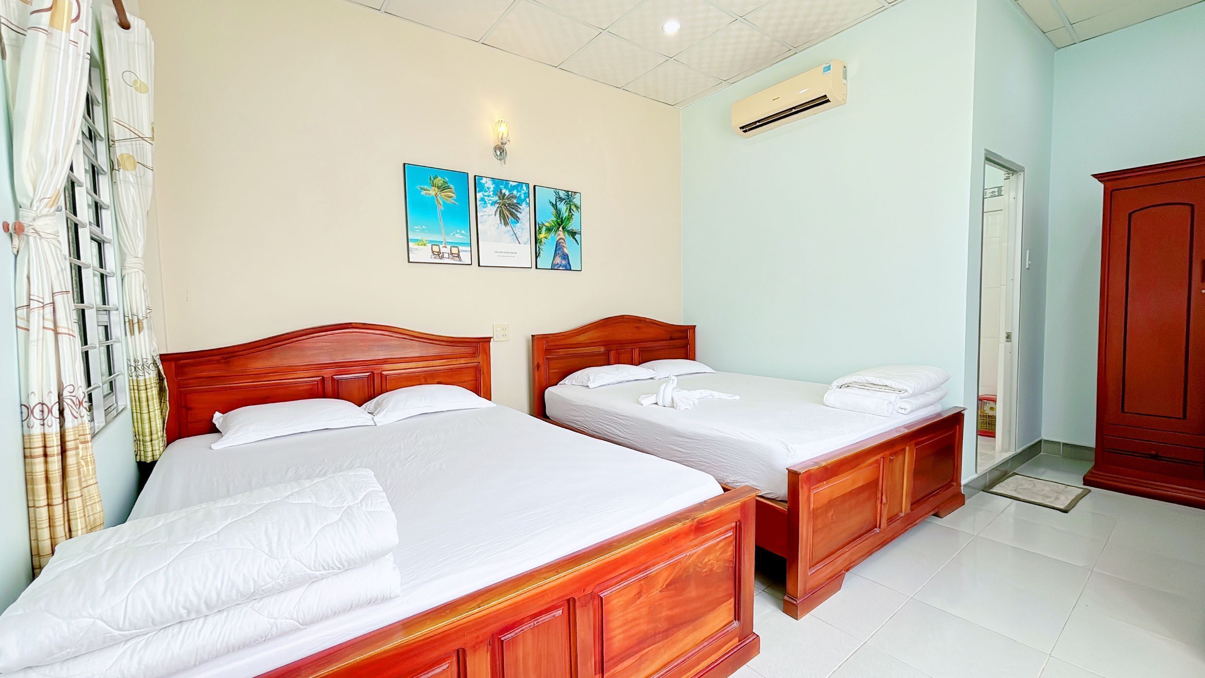 Quarto de hotel com duas camas de madeira, roupa de cama branca, ar condicionado fixado na parede e arte com tema tropical.