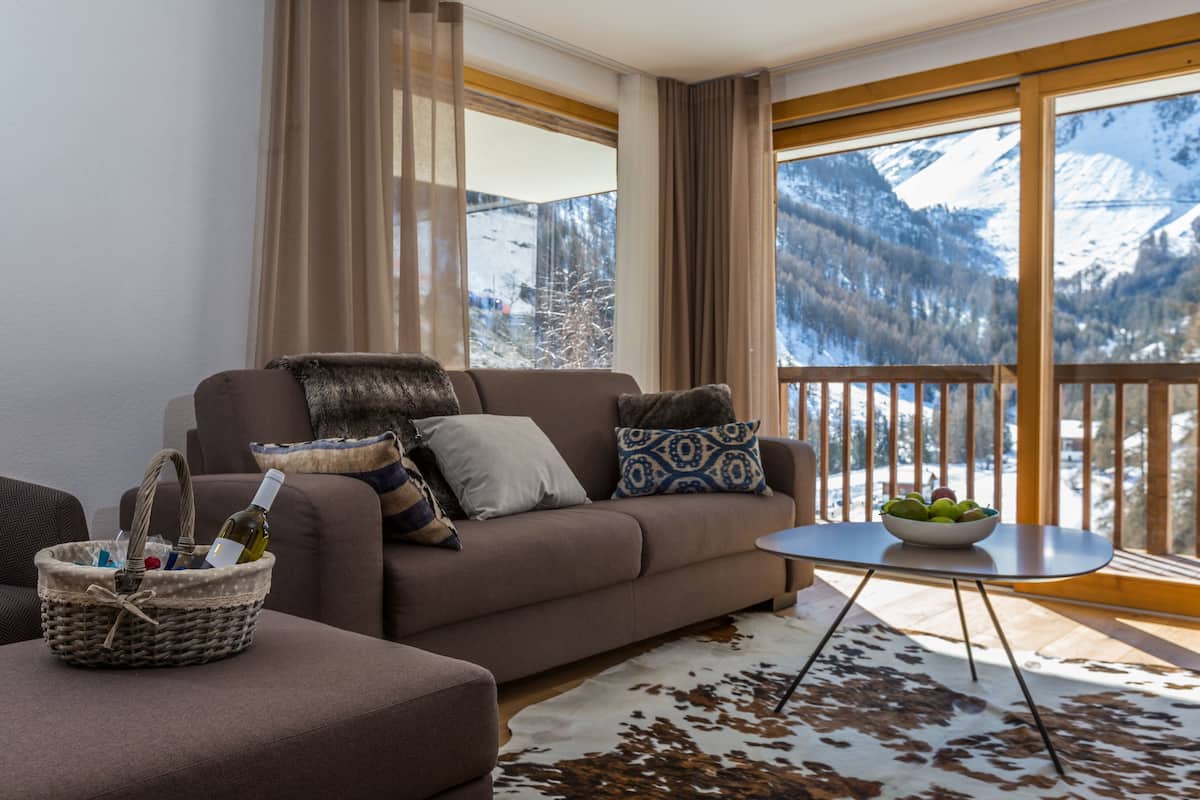 A cozy living room with a sofa, a small table, a wicker basket, and a view of snow-capped mountains through large windows.