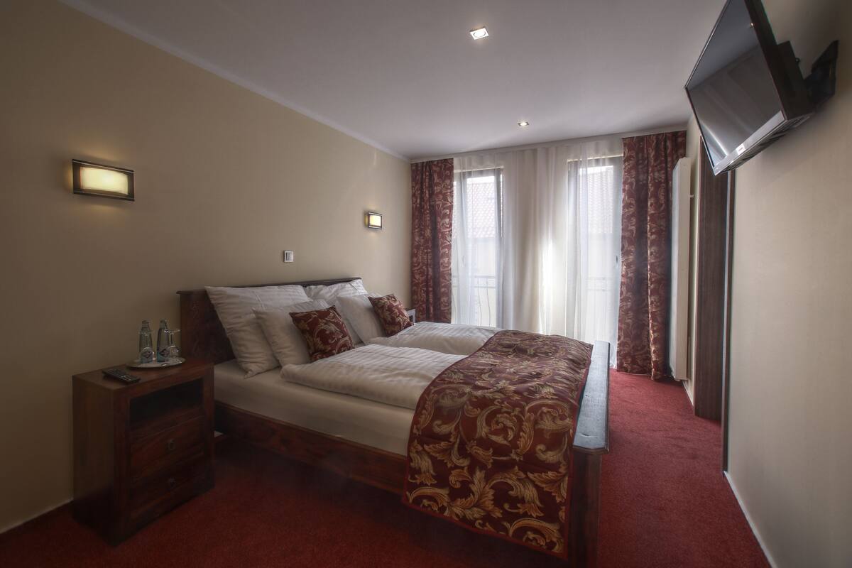 Una habitación de hotel con una cama grande, una mesita de noche con un control remoto, un televisor de pantalla plana en la pared y una ventana con cortinas.