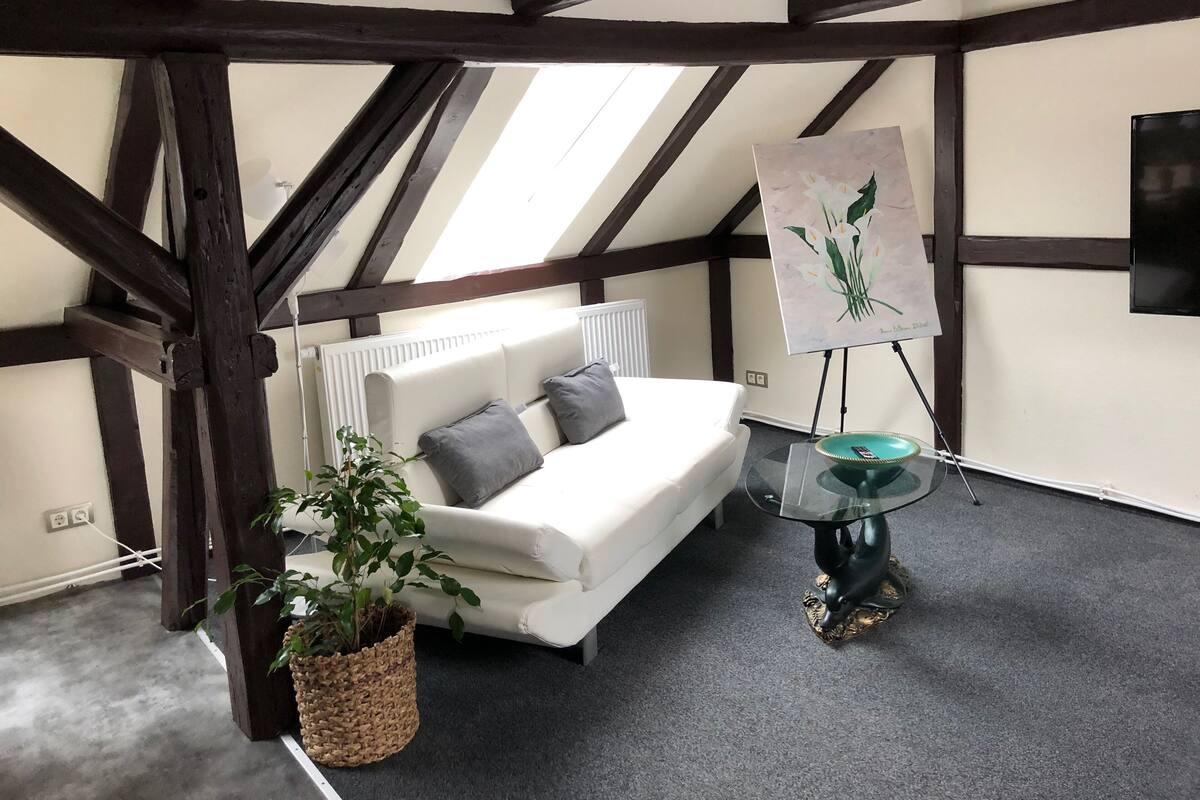 Una habitación con un sofá blanco, una planta en maceta, una mesa de vidrio con base decorativa y un cuadro sobre caballete.