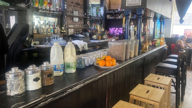 A bar area with shelves of bottles, a counter with milk cartons, and a sign above the bar. - Phoenix Hostel (London)