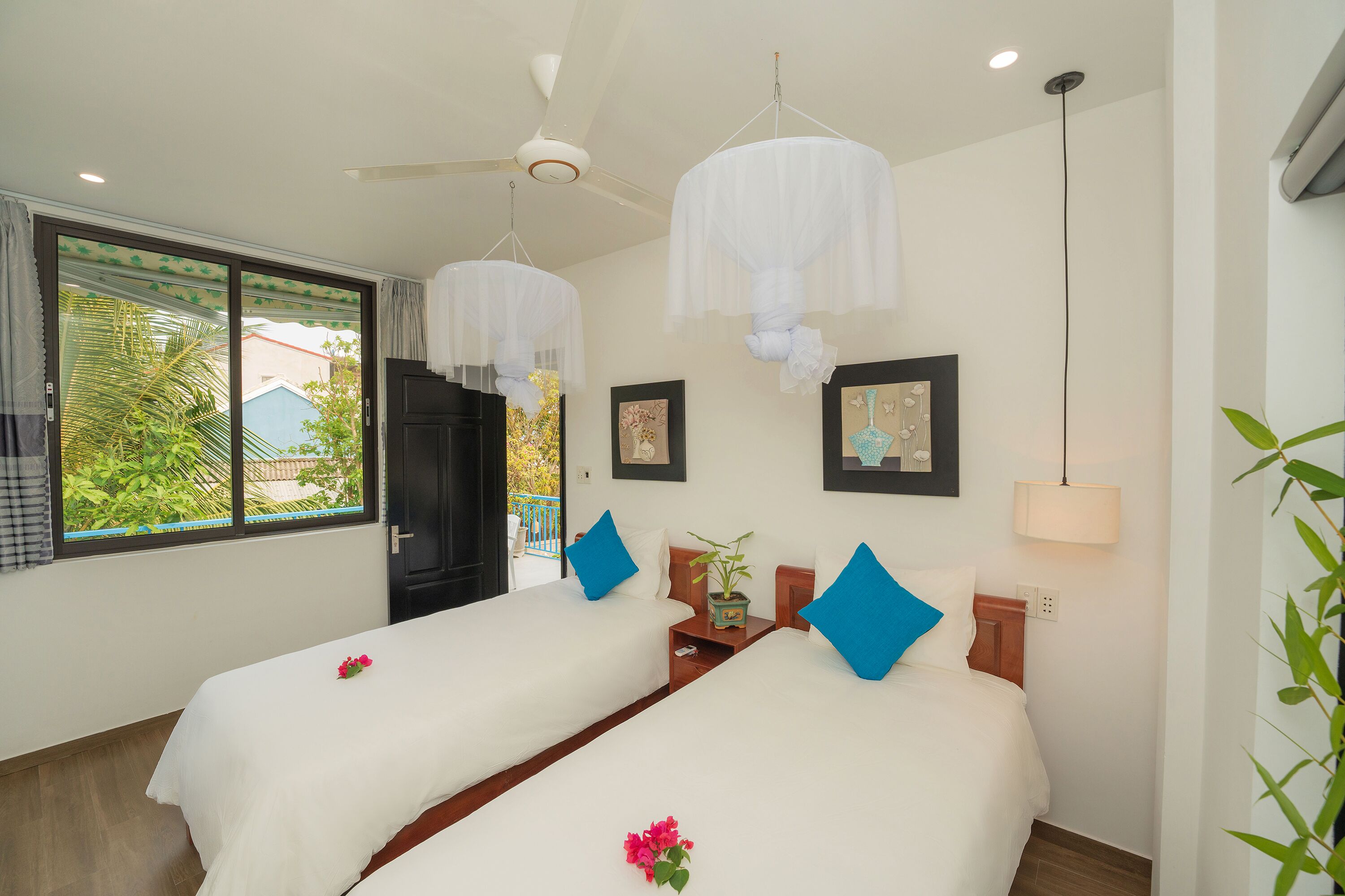 A hotel room with two beds, a ceiling fan, a window with a view of greenery, and a wall-mounted lamp.